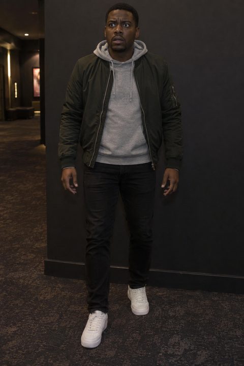 The plain capture before the poster: the same man in a bomber jacket, standing against a dark wall in a cinema corridor.